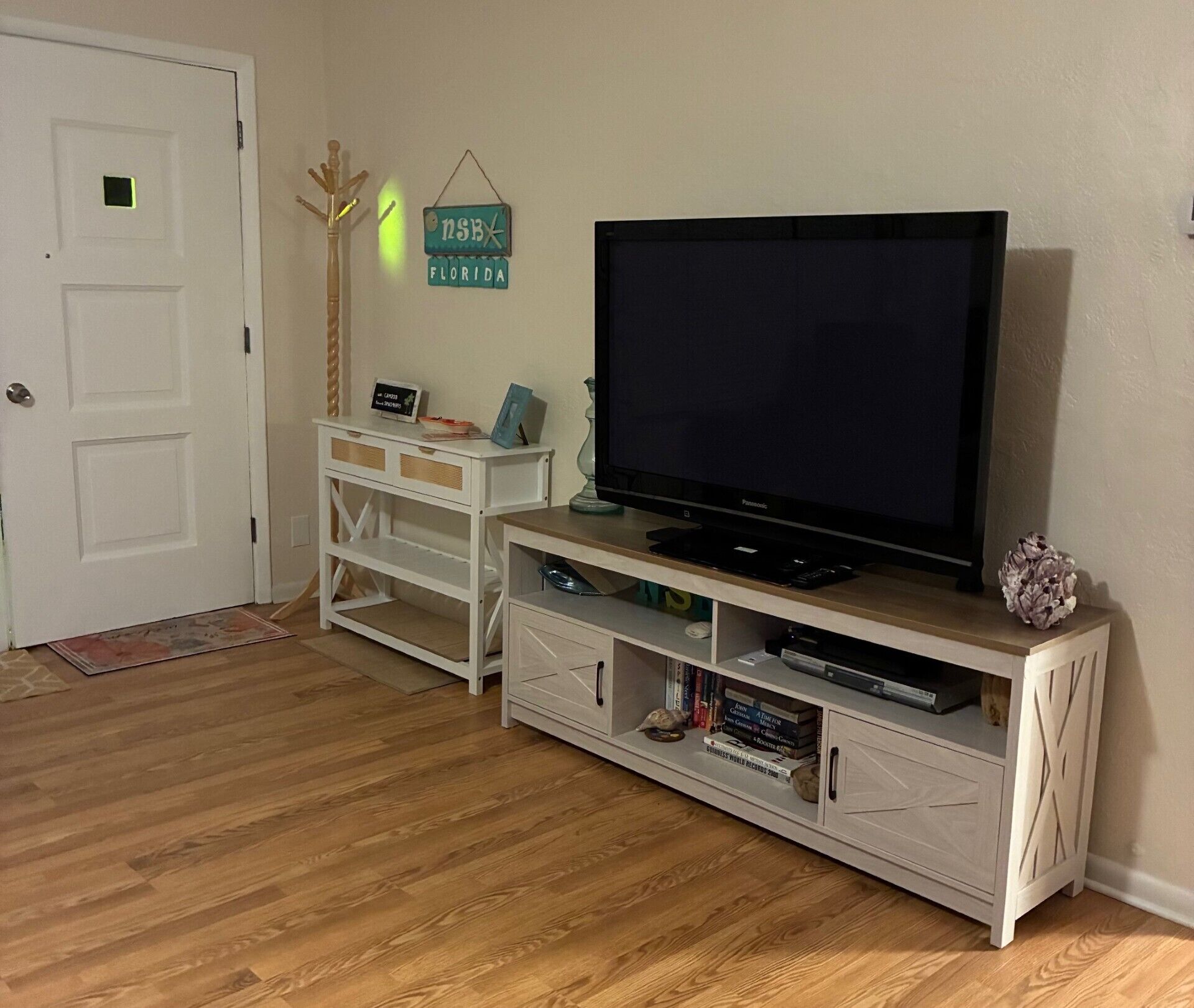 Ein Wohnzimmer mit einem Flachbildfernseher auf einer weißen Unterhaltungsanlage, Holzboden und einer Tür mit einem kleinen grünen Schild.