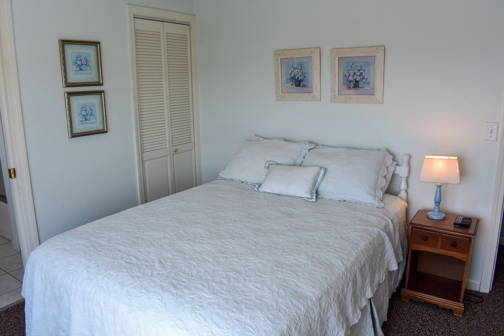 A neatly made bed with white bedding, a bedside table with a lamp, and two framed pictures on the wall.