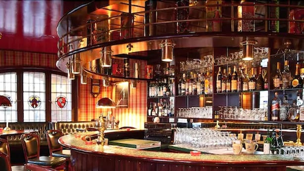 Een bar met een gebogen toonbank, verschillende zithoeken en een diverse selectie flessen en glazen op planken.