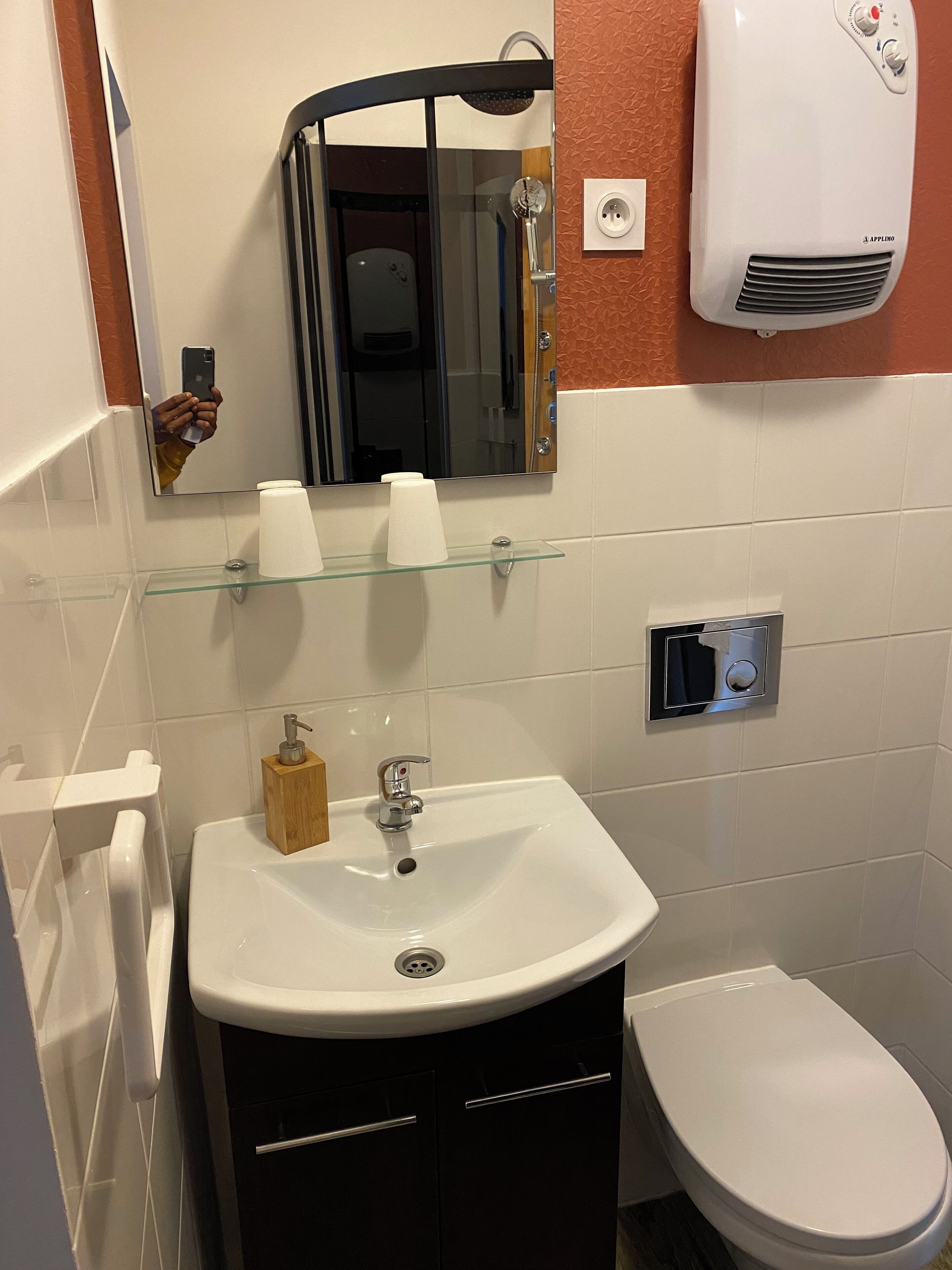 Ein Badezimmer mit einem weißen Waschbecken, einem Spiegel, einer Glasablage mit Pflegeprodukten, einer Toilette und einem an der Wand montierten Luftreiniger.