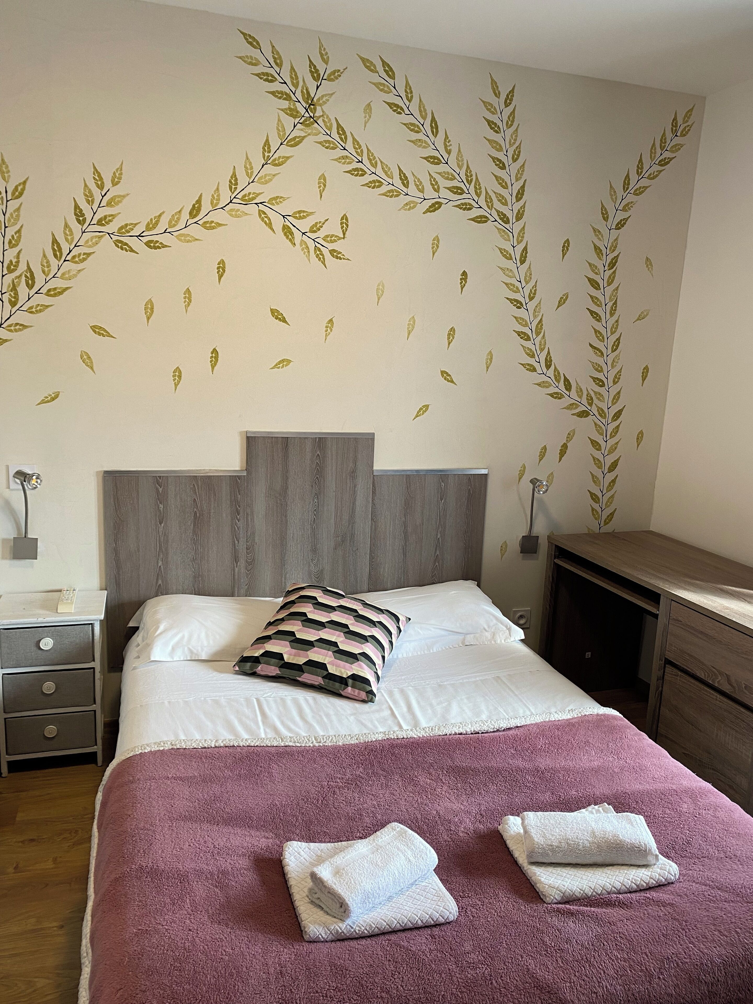 A neatly made bed with a pink blanket and two folded towels, a wooden headboard, a bedside table with a lamp, and a wall with leaf decals.
