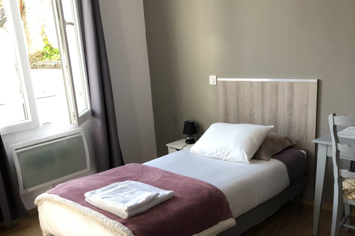 Una cama bien hecha con una manta blanca y granate, una mesita de noche con una lámpara y una silla.