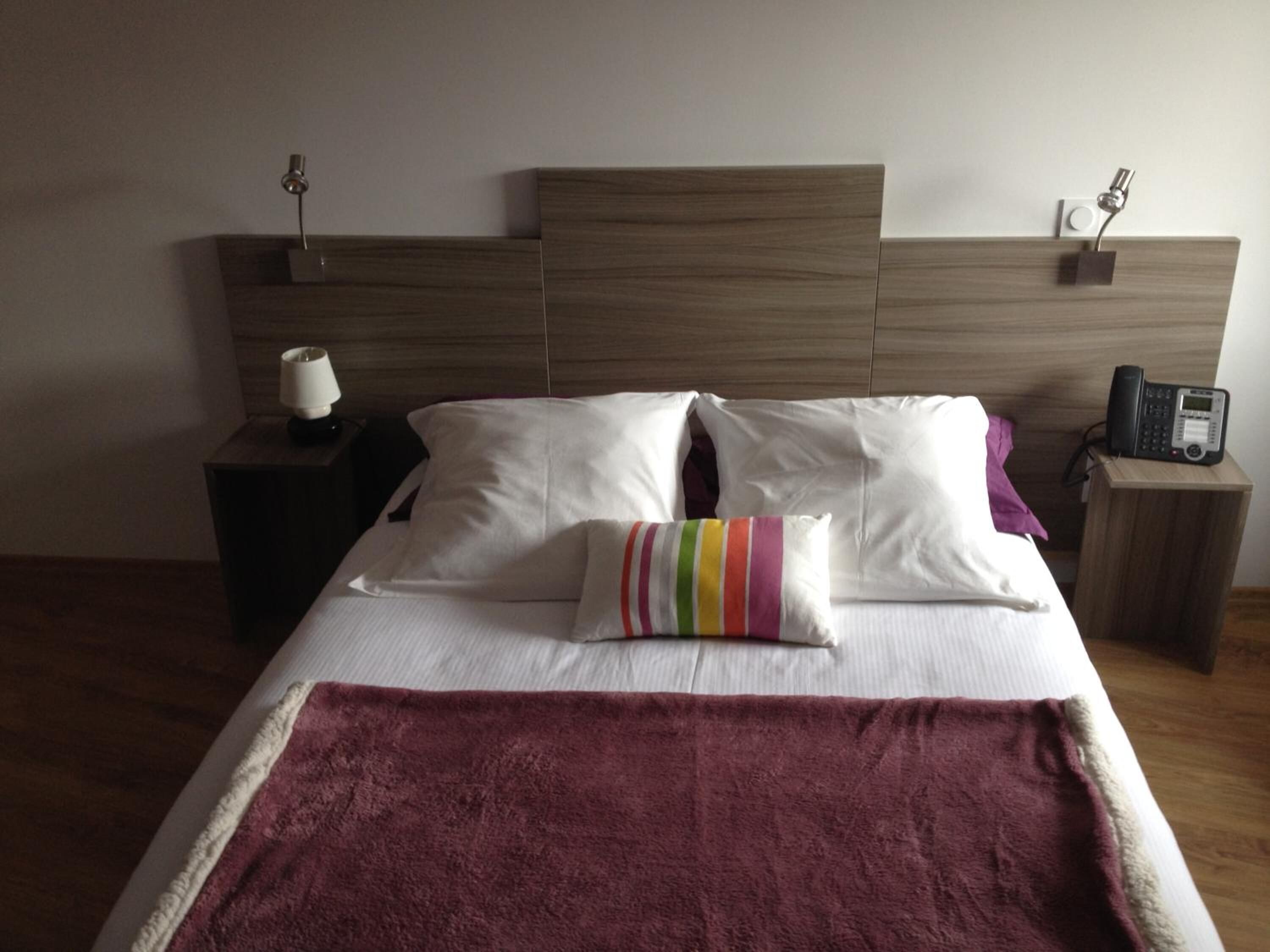 Une chambre d’hôtel avec une tête de lit en bois, un lit avec des oreillers blancs et à rayures, un plaid rouge et une table de chevet avec une lampe et un téléphone.