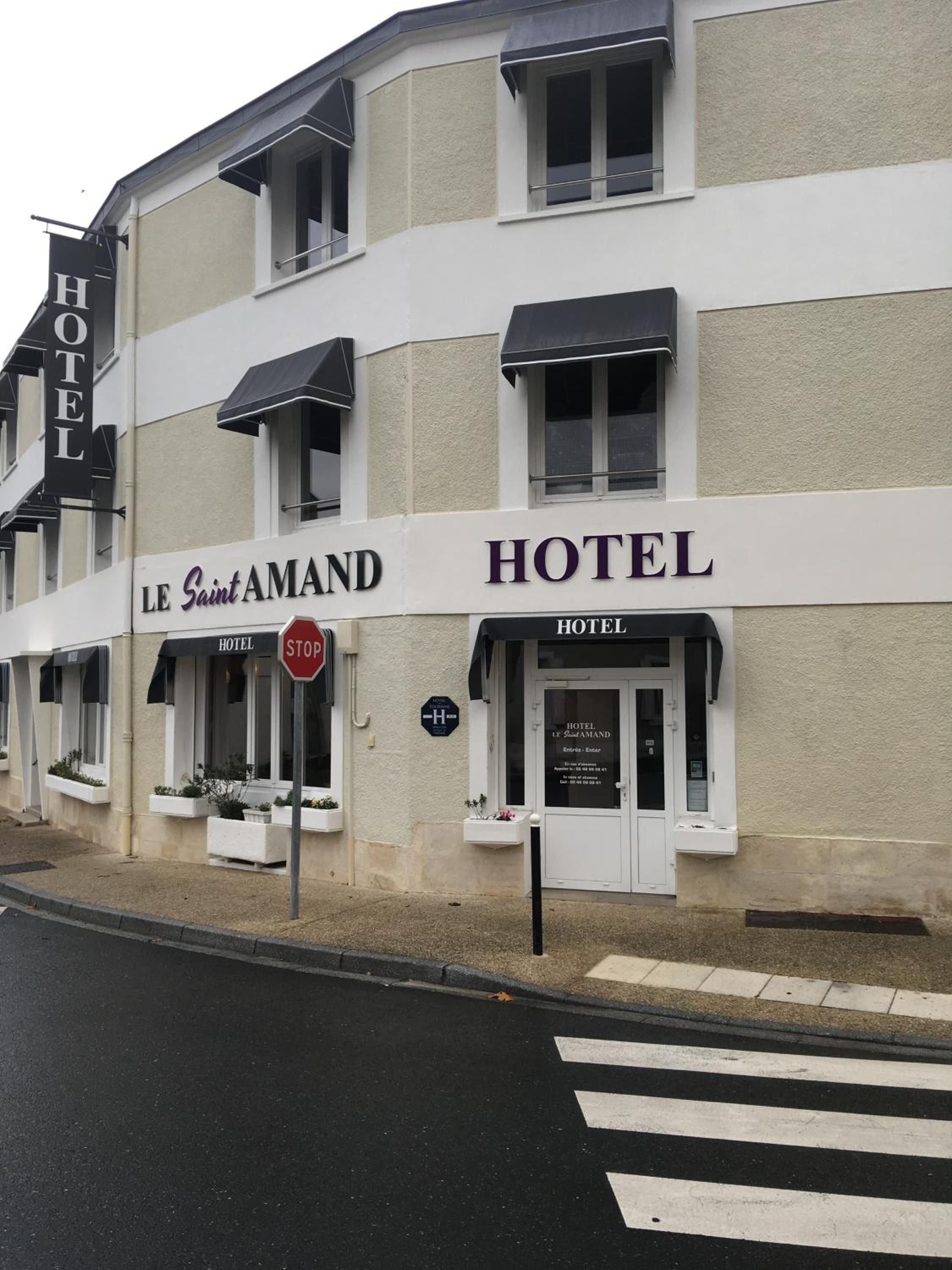 Un hôtel appelé « Le Saint Amand », avec un panneau de signalisation indiquant un arrêt juste devant, un passage piéton et une vue dégagée sur l’entrée.