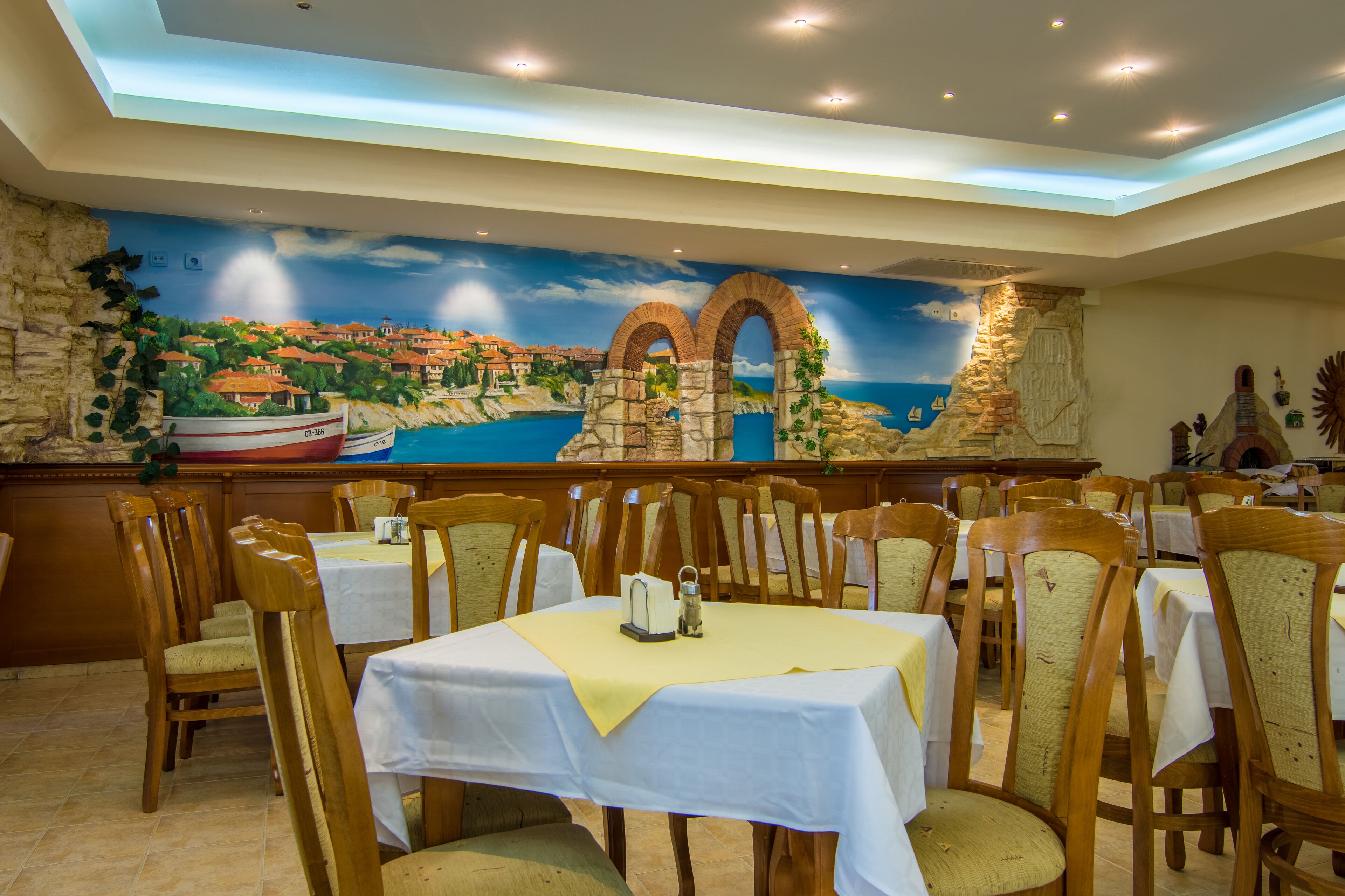 Ein Restaurant mit Wandgemälde einer Küstenstadt, Holzsesseln und Tischen mit weißen Tischdecken.