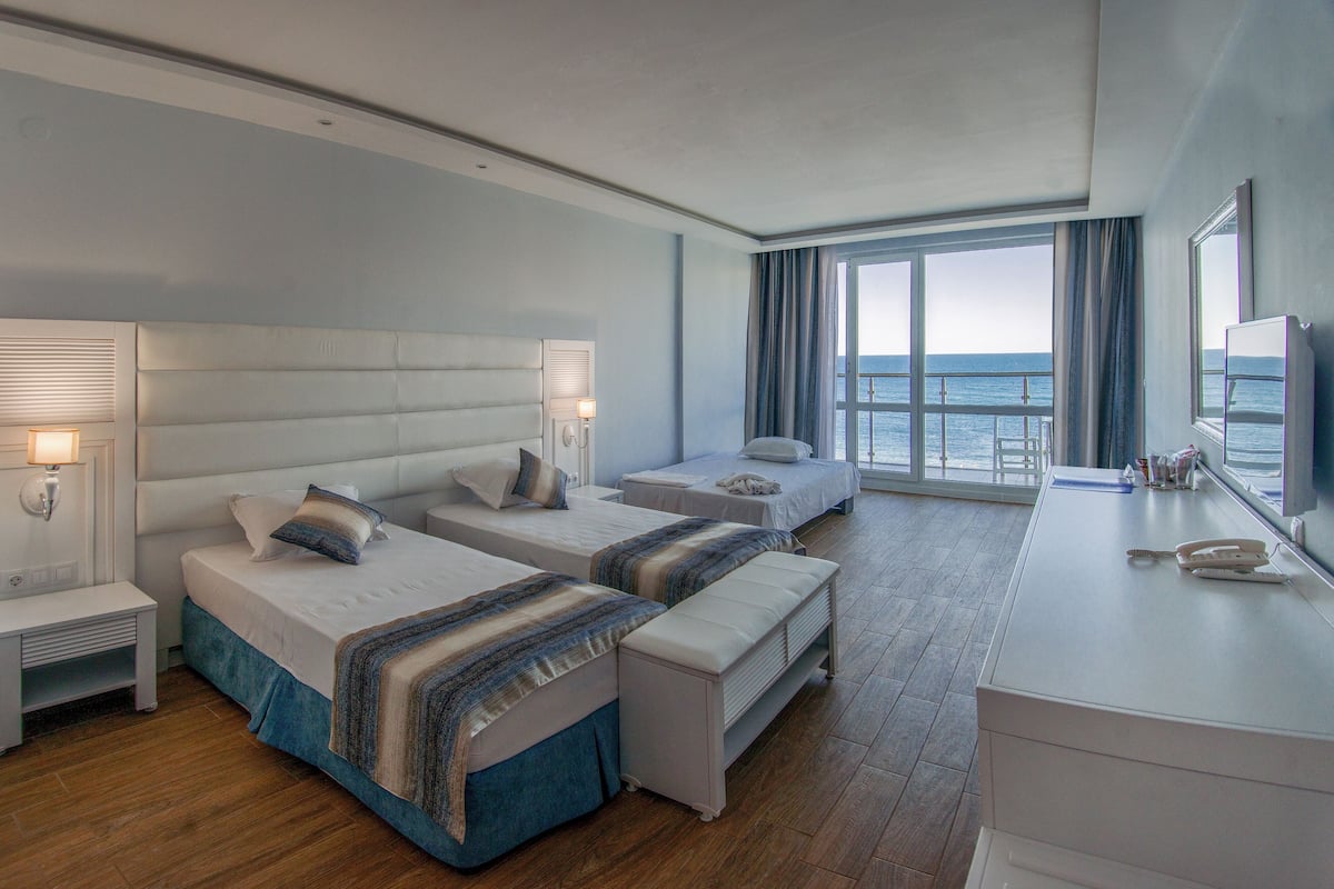A hotel room with two beds, a desk, and a large window offering a view of the sea.