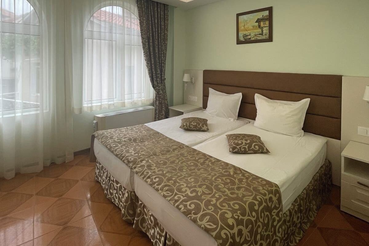 A hotel room with a large bed, two bedside tables, a nightstand, a window with curtains, and a painting on the wall.