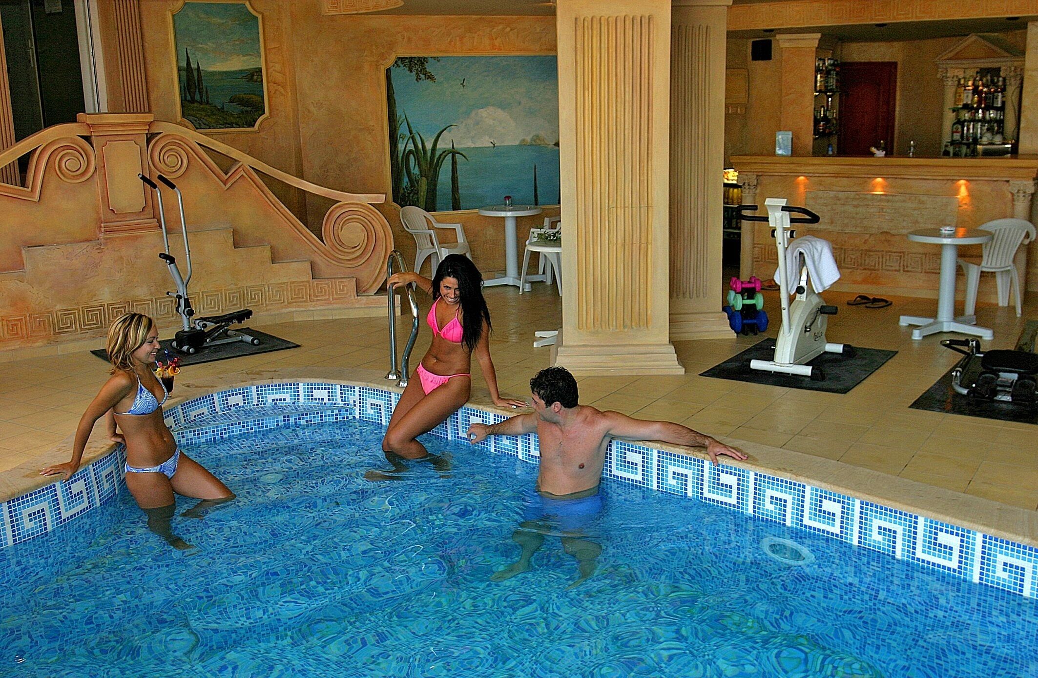 A swimming pool area with exercise equipment, a bar, and a mural on the wall.