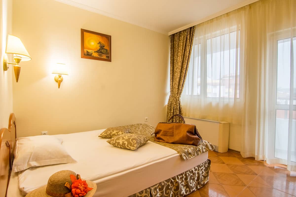 A hotel room with a bed, a wall-mounted lamp, a painting, and a window with curtains.