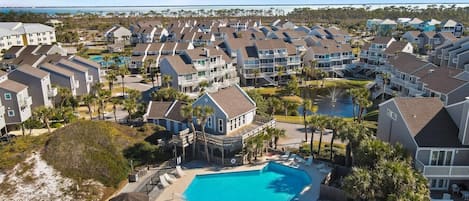 A coastal residential area with a central swimming pool, multiple-story houses, and a beach in the background.