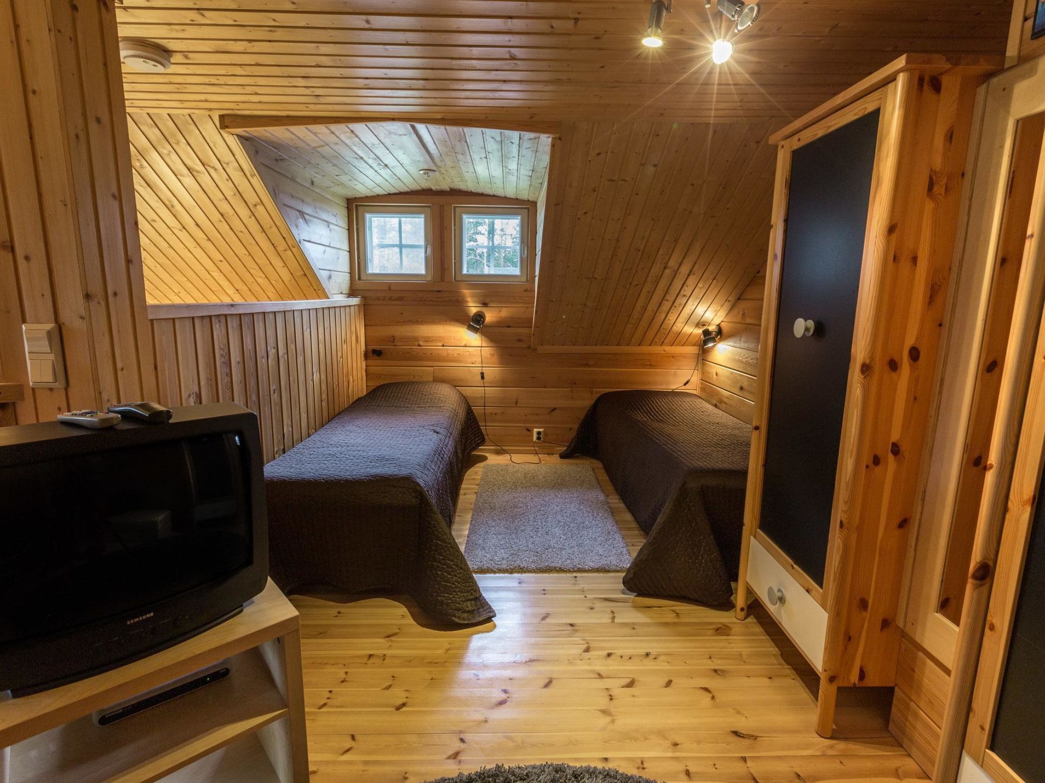 A cozy cabin room with wooden walls, a sloped ceiling, a television, two beds, and a window.