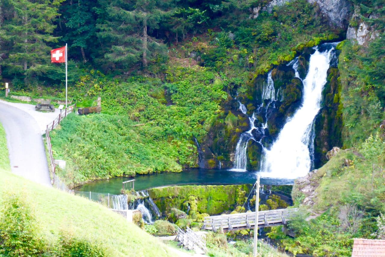 Ein Wasserfall mit Schweizer Flagge, umgeben von üppiger Vegetation und einer kleinen Brücke.