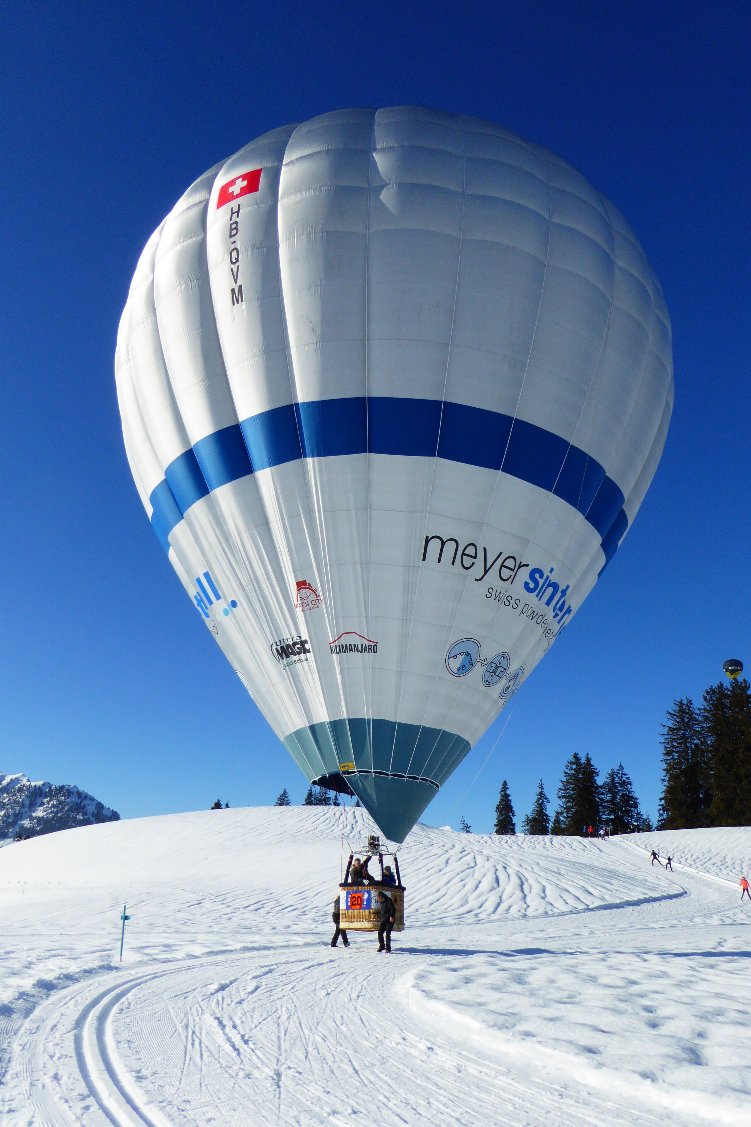 A hot air balloon with "meyersint" and "HBO-OM" logos, descending on a snowy slope.