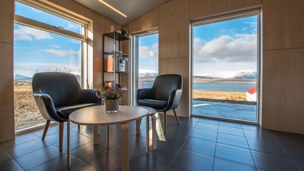 Une pièce moderne avec deux fauteuils en cuir noir, une table ronde en bois et une bibliothèque. De grandes fenêtres offrent une vue sur un lac et des montagnes.