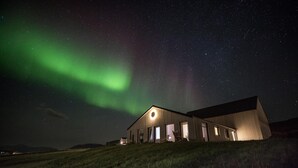 Un bâtiment moderne doté de grandes fenêtres et d’un élément lumineux circulaire sur la façade, se détachant sur un ciel nocturne parsemé d’étoiles et illuminé d’une aurore boréale verdoyante.