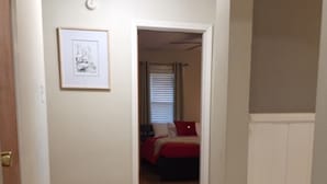 En hall med trägolv som leder till ett sovrum med ett rött sängöverkast och en inramad tavla på väggen.