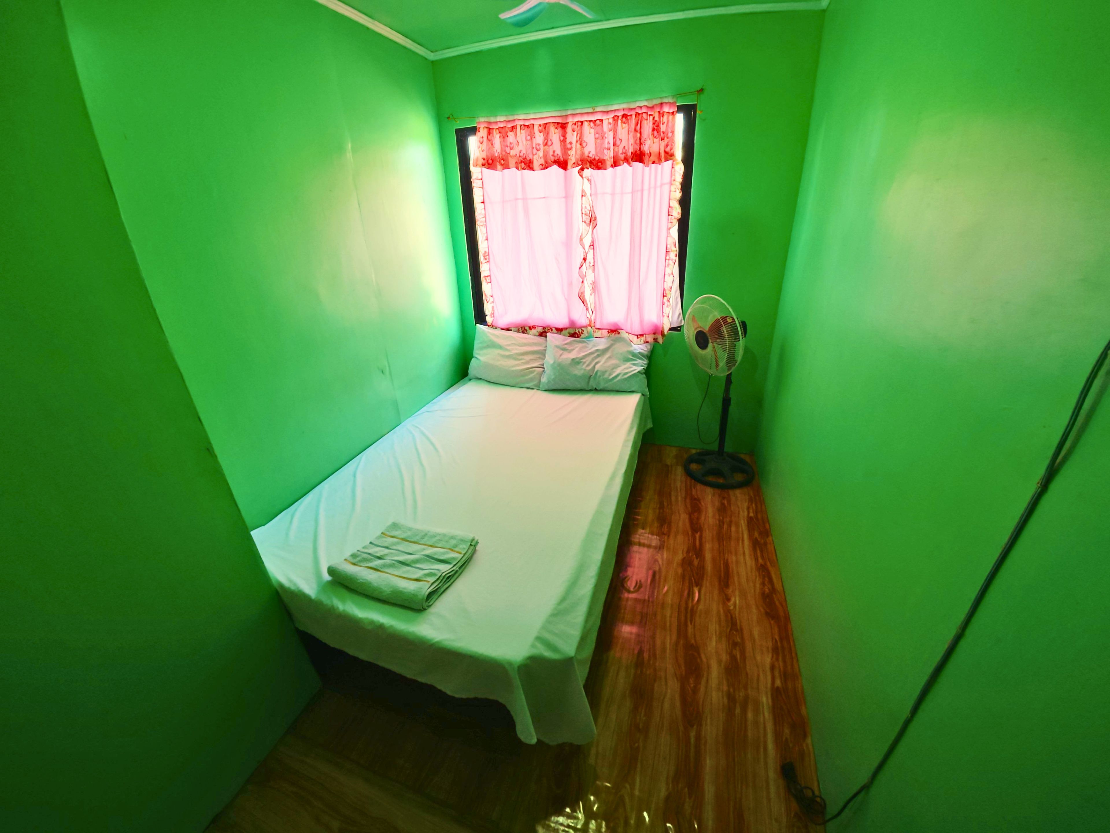 A small room with a single bed, a window with a red curtain, and a standing fan.