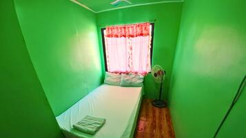 A small room with a single bed, a window with a red curtain, and a standing fan.
