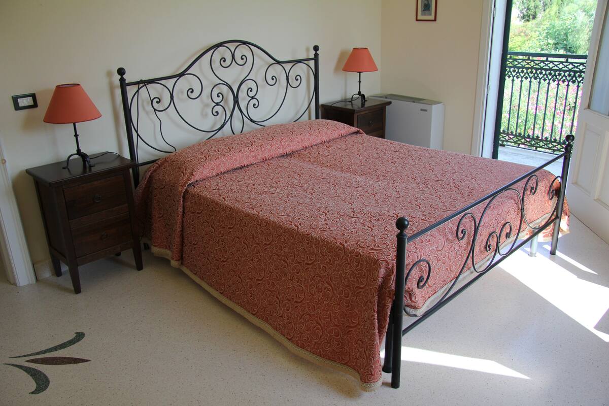 A neatly made bed with a patterned bedspread, two bedside tables with lamps, and a view of a balcony through an open door.