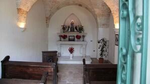 An interior with a stone archway leading to a niche with a statue, a white altar, and red flowers. There are wooden pews in front. - Parco degli Aranci (Cutrofiano)
