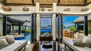 A luxurious room with a large window offering a view of a beach and ocean. There is a pool, outdoor seating, and a dining area visible through the glass doors. - WAVI Paradise, Private Luxury Island - 1 to 6 guests (Savusavu)