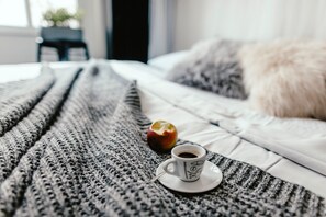 A bed with a white comforter and a gray knitted blanket. There is a white cup with a saucer and a spoon on the blanket. An apple is placed beside the cup. - Wake Up Wellness Hostel by goodnite cz (Brno)