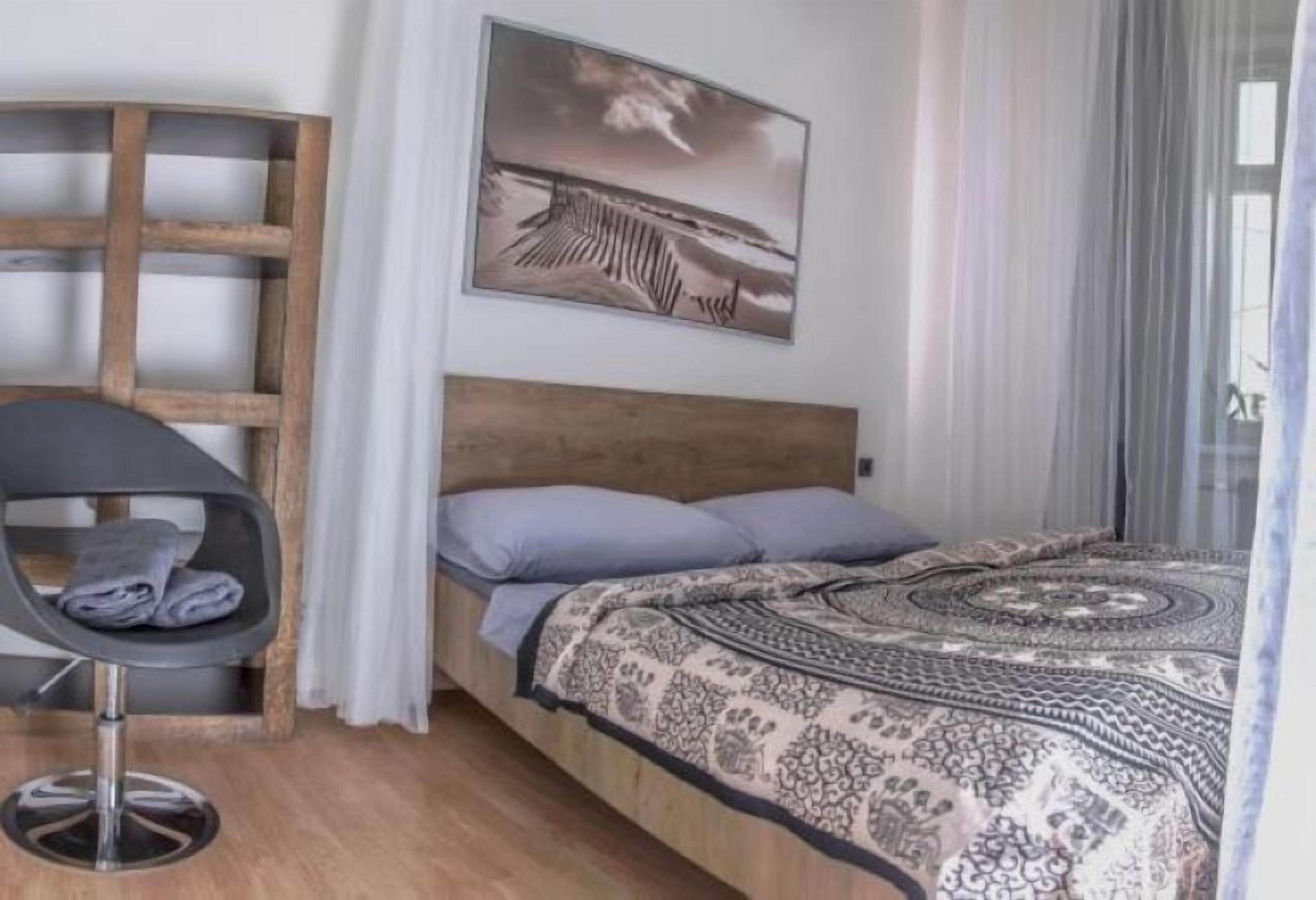 Un dormitorio con una cama, una silla, cabecera de madera, un cuadro y una ventana con cortinas.