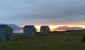 Three wooden cabins in a grassy field with a mountain and sunset in the background.