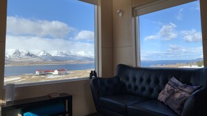 A room with a large window offering a view of a snowy mountain range and a body of water. - Sunnuhlid houses (Akureyri)