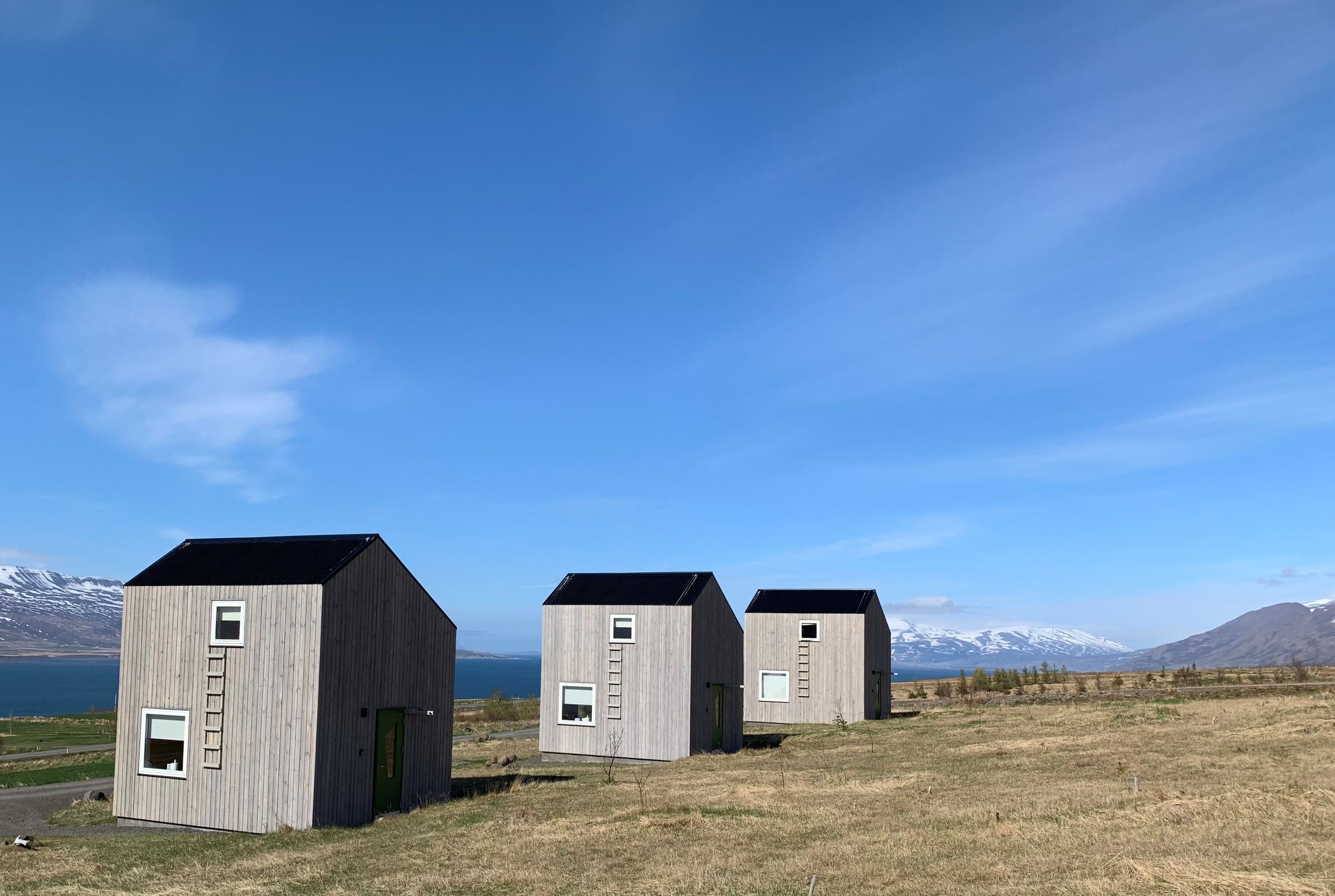 Three wooden cabins with external ladders, set against a backdrop of mountains and a lake.