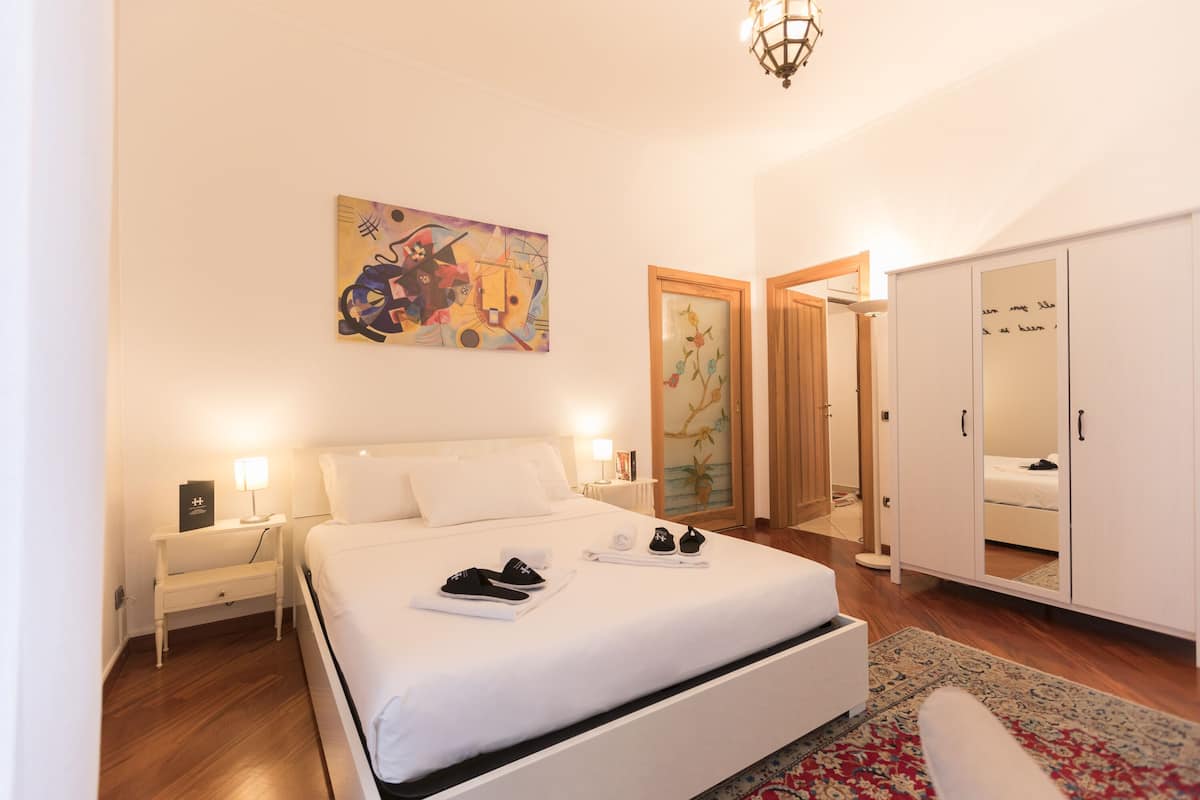 A hotel room with a large bed, a nightstand with a lamp, a wardrobe, and a painting on the wall.
