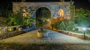 Une arche en pierre ornée de statues, dont l’intérieur est illuminé, et qui est entourée de plantes et bordée d’un chemin pavé.