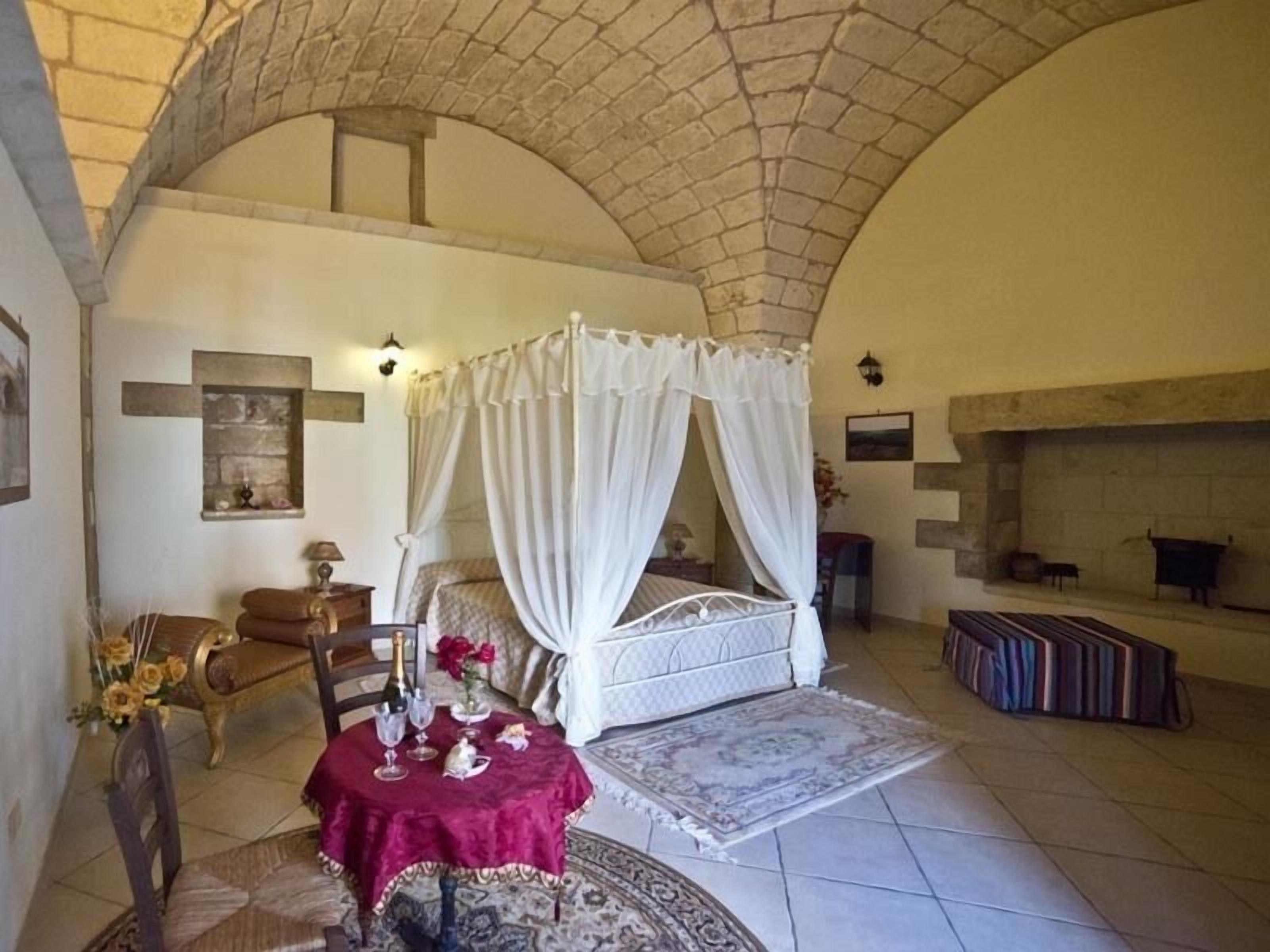 A bedroom with a four-poster bed, a small table with a red tablecloth, a chair, and a fireplace.