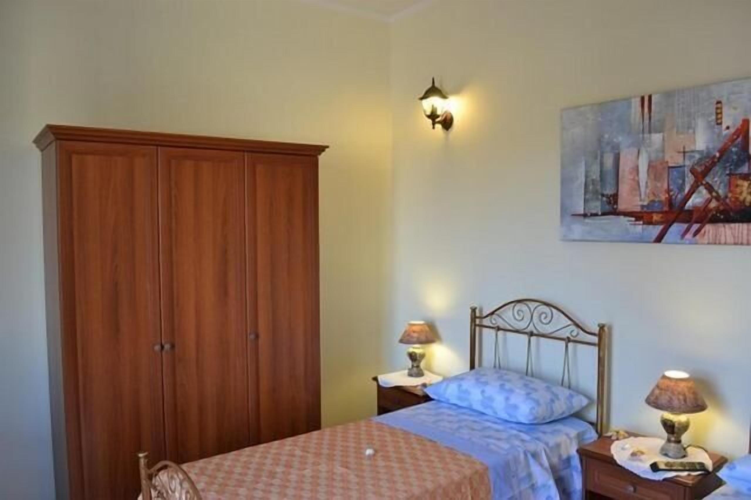 Ein Schlafzimmer mit einem Holzschrank, einem Bett mit blau-weißer Karomusterausstattung, zwei Nachttischen inklusive Lampen sowie einem Gemälde an der Wand.