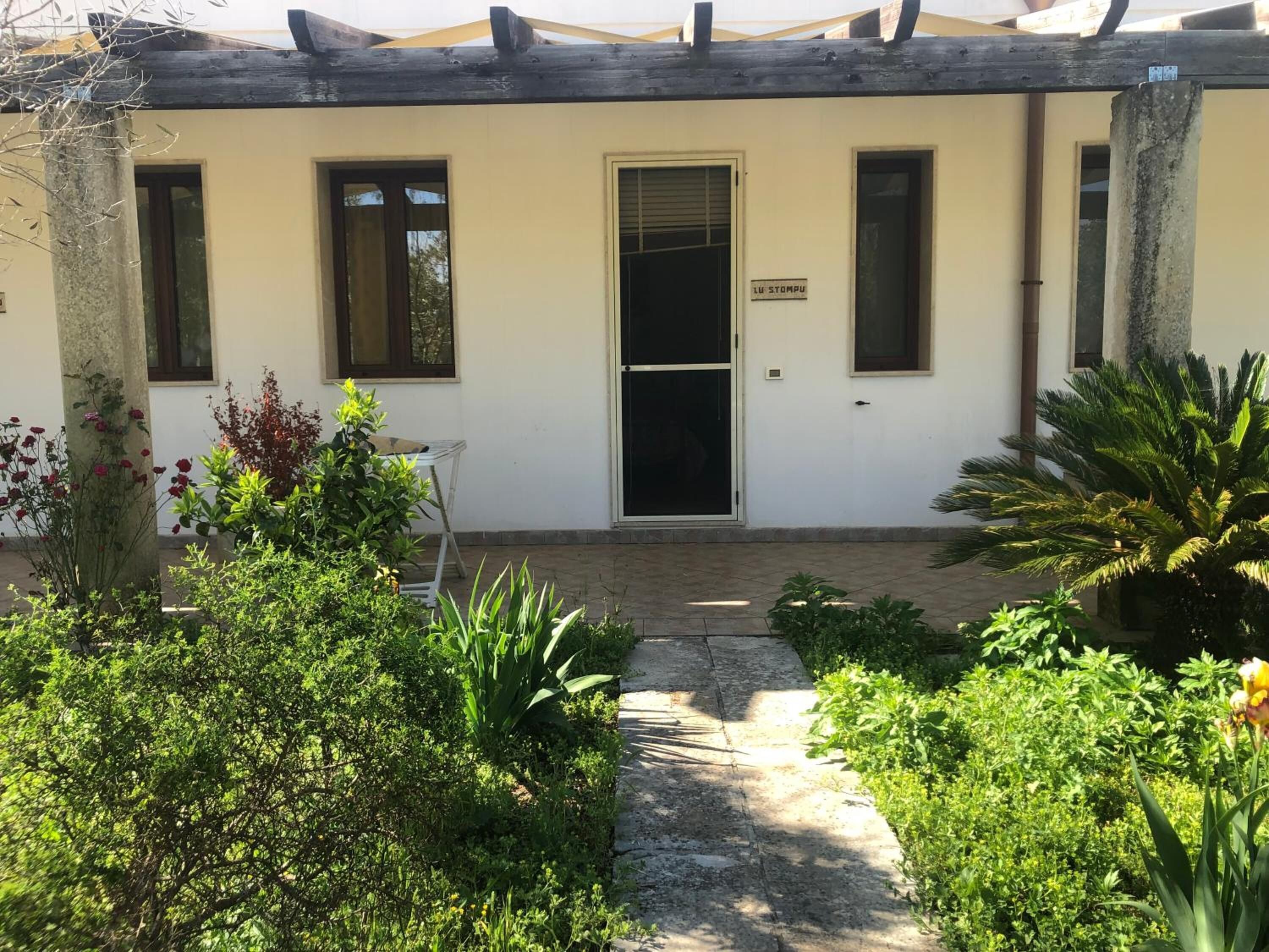 A building with a white exterior, a door with a sign, and a small garden area with various plants and a chair.