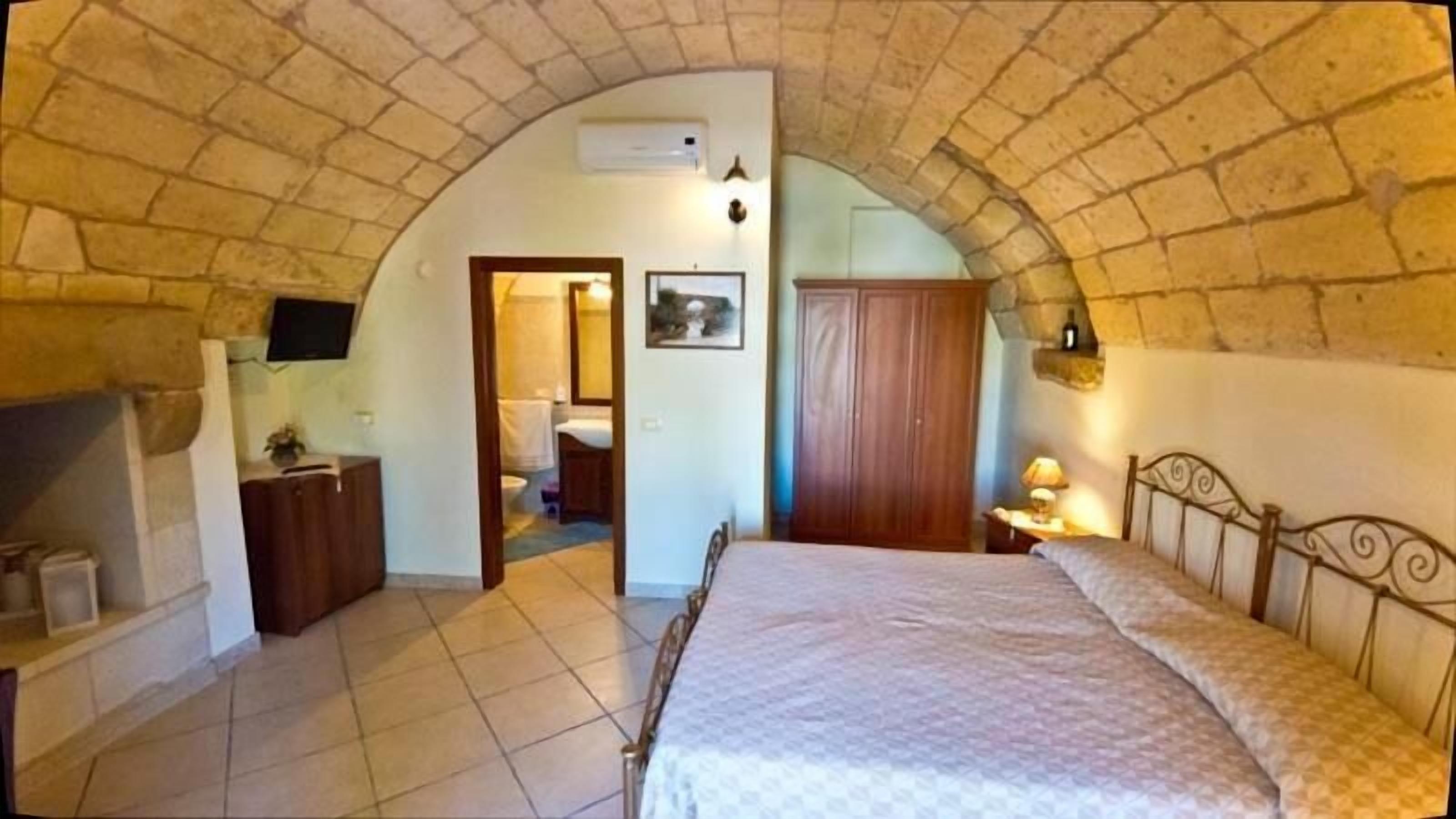 A bedroom with a stone vaulted ceiling, a bed, a wooden wardrobe, a bedside table with a lamp, and a bathroom visible through an archway.
