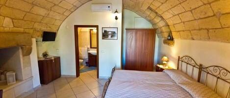 A bedroom with a stone vaulted ceiling, a bed, a wooden wardrobe, a bedside table with a lamp, and a bathroom visible through an archway.