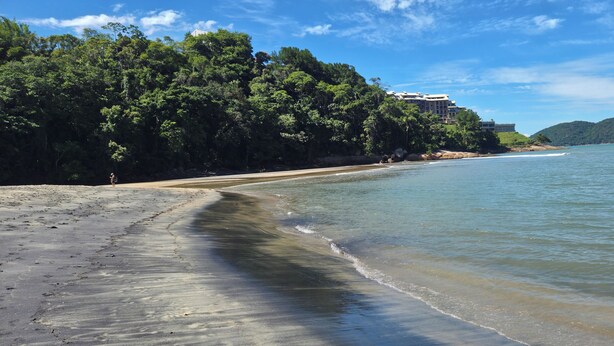 A beach with a sandy shore, a building with multiple stories, and a forested hill in the background.