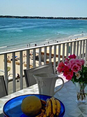 Une table avec une carafe, une assiette de fruits et un vase de fleurs, donnant sur une plage où se trouvent des gens et une mer bleue et limpide.