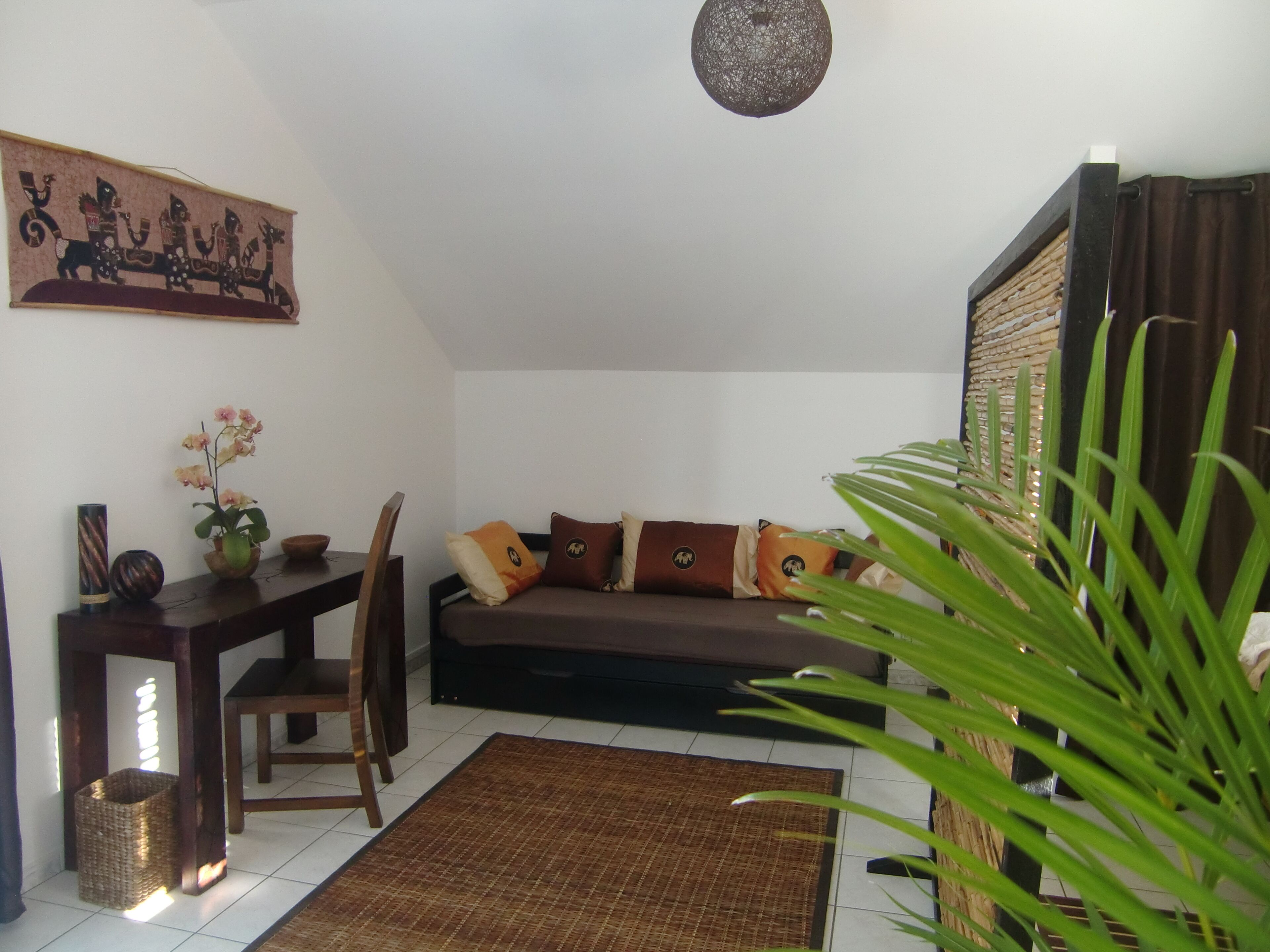 A living room with a sofa, a wooden table, a chair, a decorative wall hanging, and a large plant.