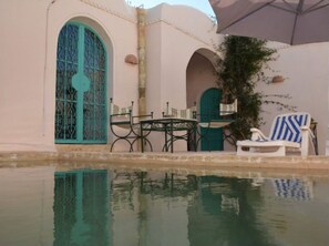 A poolside area with a table and chairs, a blue door, and a covered umbrella. - DAR LILY offers you an authentic and hidden Djerbian experience. (Djerba)