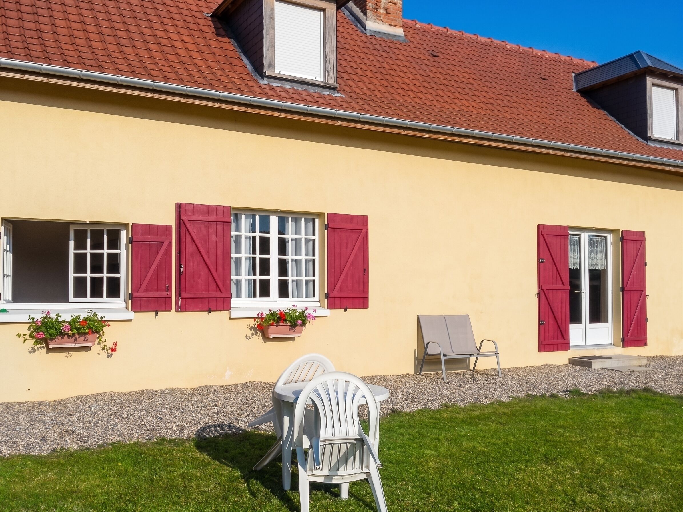 Une maison à la façade jaune, aux volets rouges et à la petite terrasse où se trouvent une chaise blanche et une table.