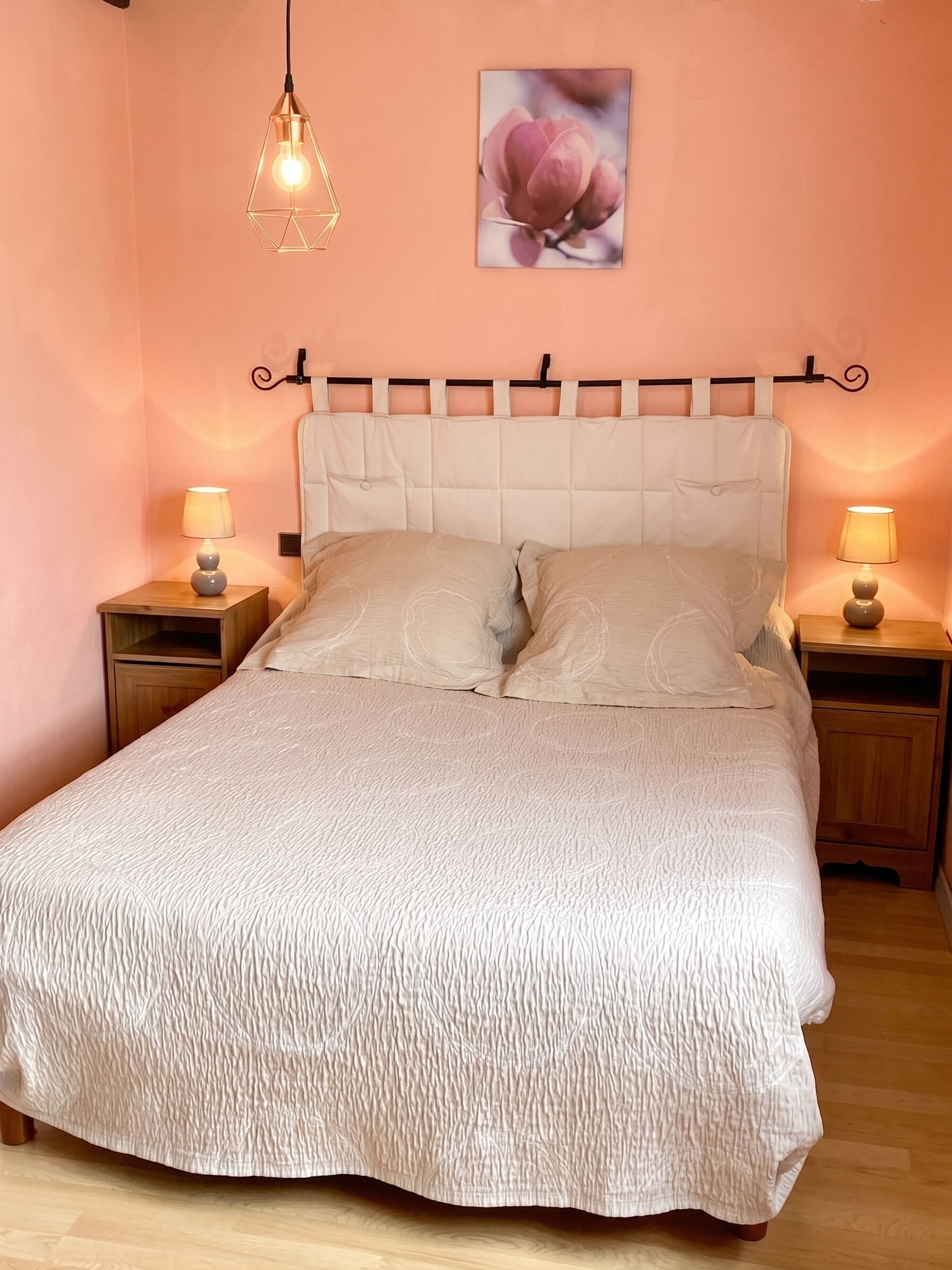 Ein Bett mit einer strukturierten, beige farbenen Bettdecke, zwei Nachttischlampen, ein an der Wand befestigtes Bild von einer Blume und eine geometrische Pendelleuchte.