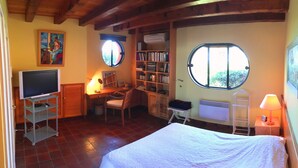 Ein Schlafzimmer mit einem Bett, einem Schreibtisch mit Stuhl, einem Fernseher auf einem Schrank, einem Bücherregal und zwei Fenstern.