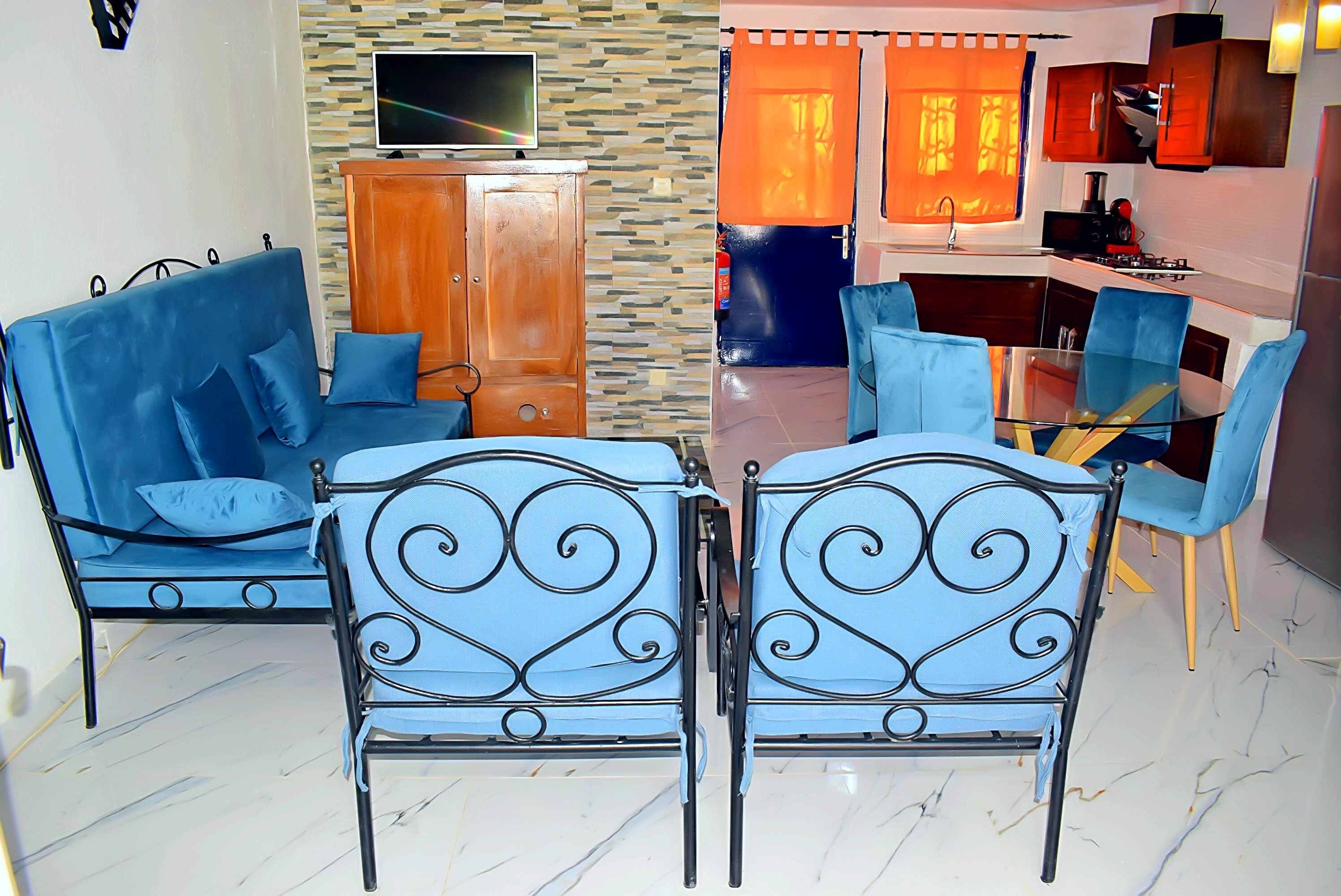 Un salon comprenant un canapé, deux fauteuils, un téléviseur et un coin cuisine avec une table et des chaises.