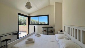 Une chambre moderne avec un grand lit, une petite table de chevet, une commode, une fenêtre donnant sur un espace verdoyant et un luminaire suspendu.