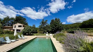Une piscine rectangulaire à l’eau claire, entourée d’une terrasse bordée de galets. On y trouve plusieurs chaises longues et un parasol blanc. En arrière-plan, on aperçoit une maison avec un toit en pente, entourée d’arbres et d’un paysage vallonné.