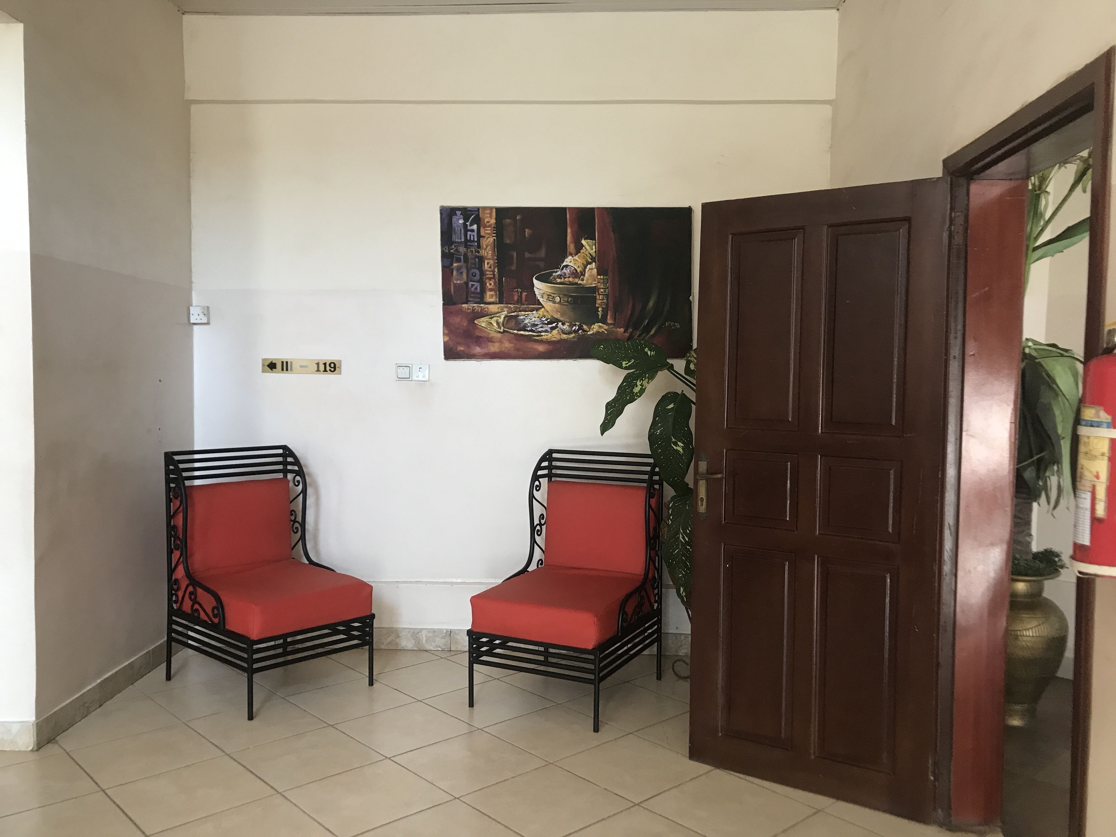A hallway with two red cushioned chairs, a wooden door, a wall-mounted painting, and a potted plant.