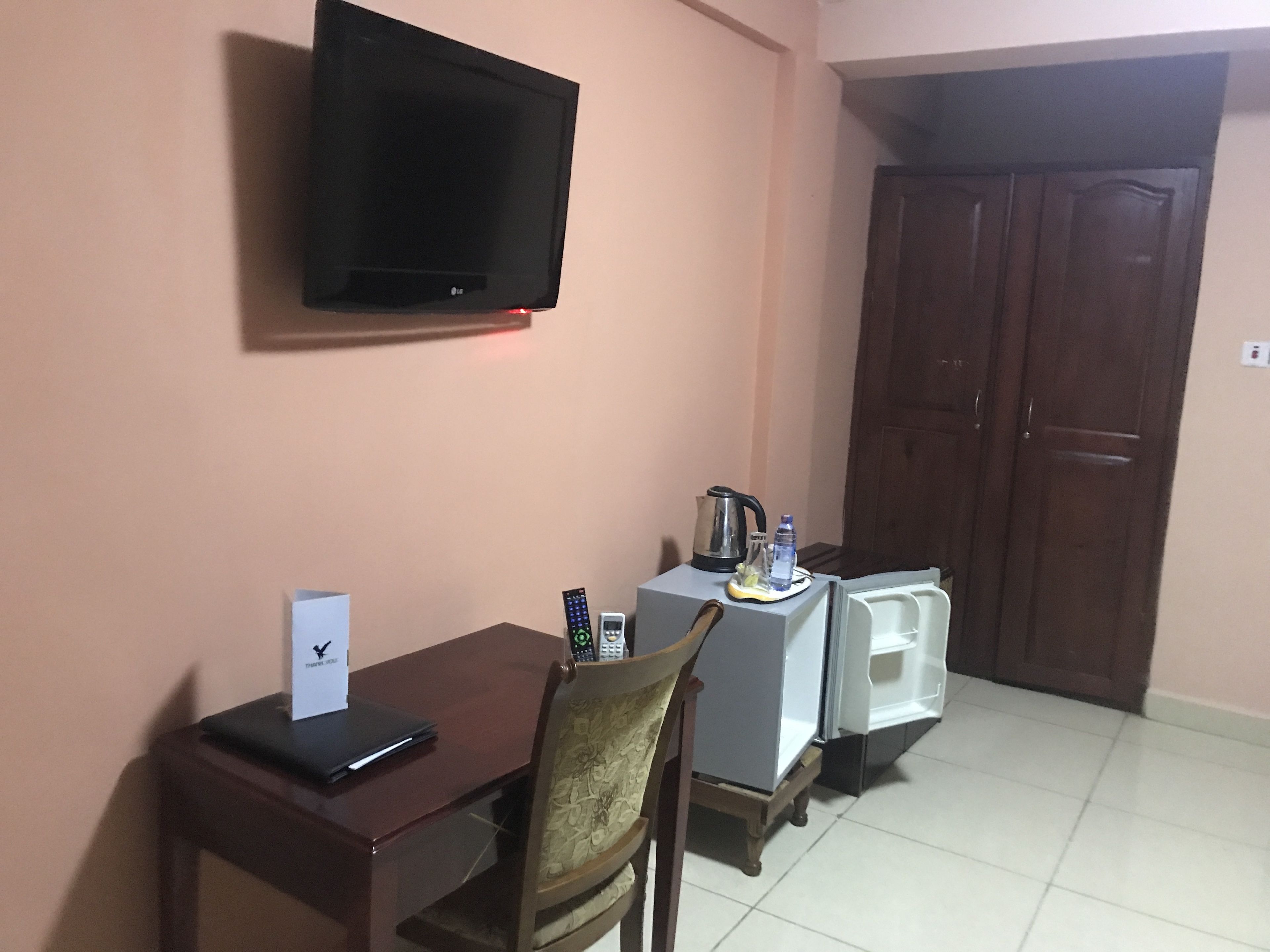 A hotel room with a television mounted on the wall, a wooden table with a chair, a small refrigerator, and a cabinet.