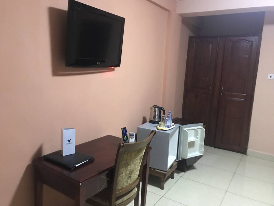 Une chambre d’hôtel équipée d’un téléviseur fixé au mur, d’une table en bois avec une chaise, d’un petit réfrigérateur et d’un placard.
